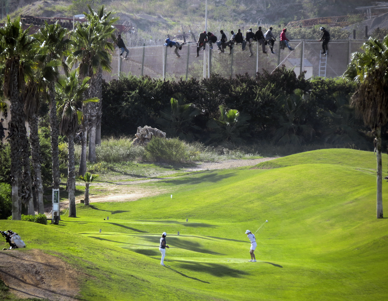 A golfer hits a tee shot as African migrants sit atop a border fence during an attempt to cross into Spanish territories between Morocco and Spain's north African enclave of Melilla